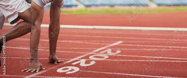 Fototapeta A runner crouches at the starting line of a track, getting ready to sprint. The clean red surface and numbered lanes reveal an active sports environment on a bright day.