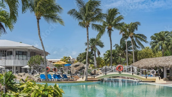 Fototapeta Swimming pool in a tropical resort. Arched pedestrian bridge over turquoise water. A lifebuoy on a rope railing. A canopy with a thatched roof. Palm trees against the blue sky. Cuba. Varadero. Hotel