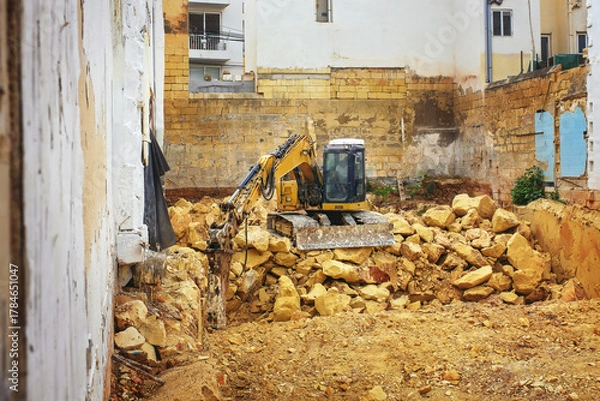 Obraz Industrial excavator breaking rocks and clearing foundation pit at construction site surrounded by old stone buildings.