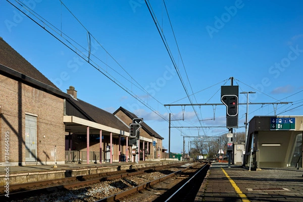 Fototapeta Bright, sunny day at a quiet train station with multiple tracks and platforms under a clear blue sky. Brick station buildings line the left, while modern elements and signal lights mark the right.