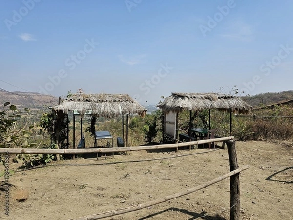 Fototapeta Countryside shelter A handmade straw-roof hut beside a dirt road, set against a scenic mountain backdrop and open horizon, ideal for nature or travel themes.