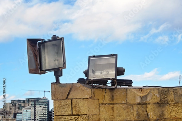 Obraz Two industrial floodlights mounted on a stone wall against a cloudy blue sky and modern city skyline on the horizon.