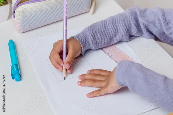 Fototapeta A little girl drawing something on paper using a pencil on the table