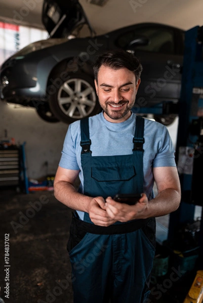 Fototapeta Mechanic checking phone while working on car in garage during the day