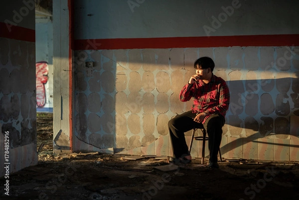 Fototapeta Young Asian man in a plaid shirt sitting and smokes a cigarette in abandoned building alone.