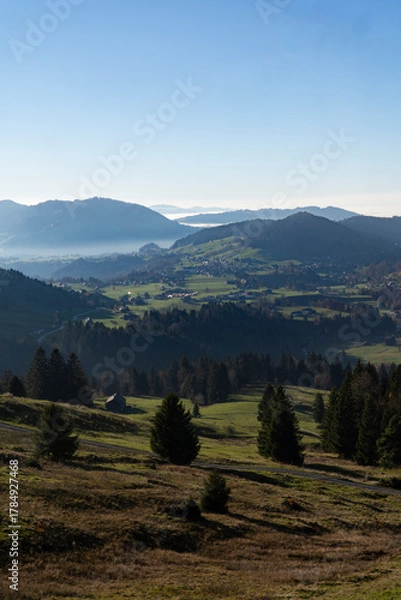Fototapeta misty but serene view mountain valley with green fields and distant hills under clear blue sky in Bregenzer Wald in Vorarlberg Austria