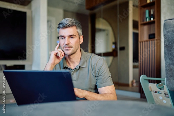 Obraz Middle-aged man sitting in his living room, working remotely on a laptop, concentrating and thinking deeply