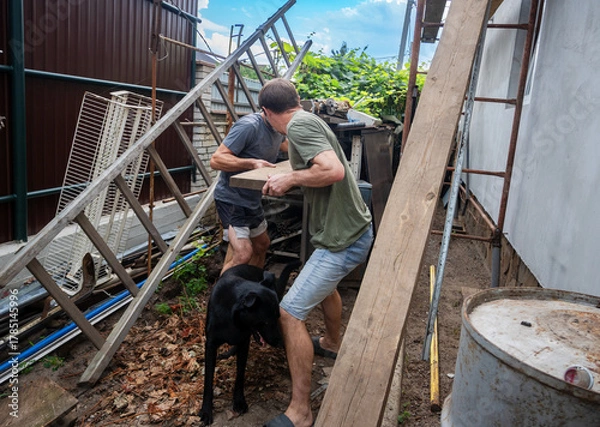 Fototapeta Two men carry a wooden plank amidst construction debris in a cluttered backyard, accompanied by a black dog. Shows teamwork and hard work during renovation or cleanup projects outdoors.