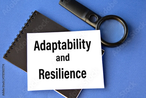 Fototapeta ADAPTABILITY and RESILIENCE - words on a white sheet on a black notebook with a magnifying glass