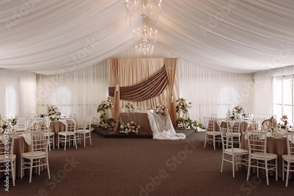 Fototapeta Elegant wedding banquet hall with draped ceiling, chandeliers, floral arrangements, and decorated stage in beige and white tones. Concept for wedding venue, celebration, luxury event, decor design.
