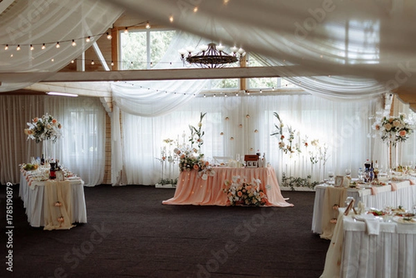 Fototapeta Beautiful wedding reception setup with white flowers, green accents, round tables, and soft lighting in a wooden hall with draped fabric ceiling. Concept: celebration, romance, event design, luxury.