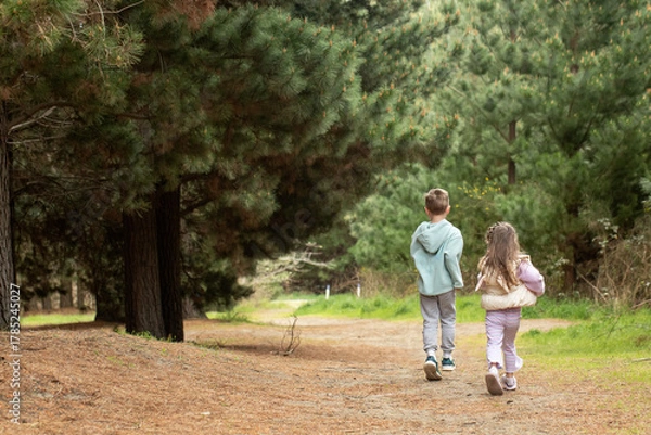 Fototapeta Two children running together along a forest path, surrounded by tall green pine trees. 