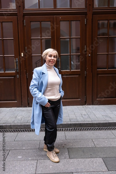 Fototapeta A blonde woman in a white sweater and scarf stands outside a business building. The architecture of the building is dominated by red brick.