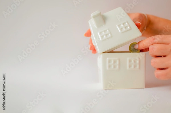 Obraz Hand dropping coin into a house-shaped piggy bank representing savings and investment