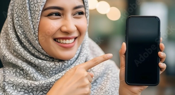 Fototapeta A cheerful young woman wearing a hijab smiling and pointing at a smartphone in a modern indoor setting