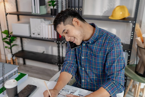 Fototapeta A young architect is multitasking at his desk, sketching blueprints, making phone calls, and reviewing notes. His workspace shows focus, productivity, and modern design work.