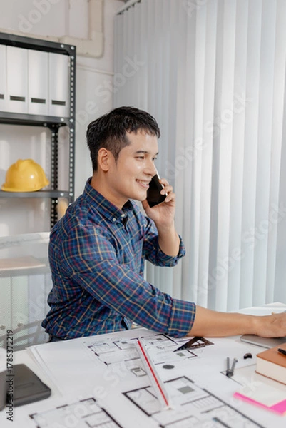 Fototapeta A young architect is multitasking at his desk, sketching blueprints, making phone calls, and reviewing notes. His workspace shows focus, productivity, and modern design work.