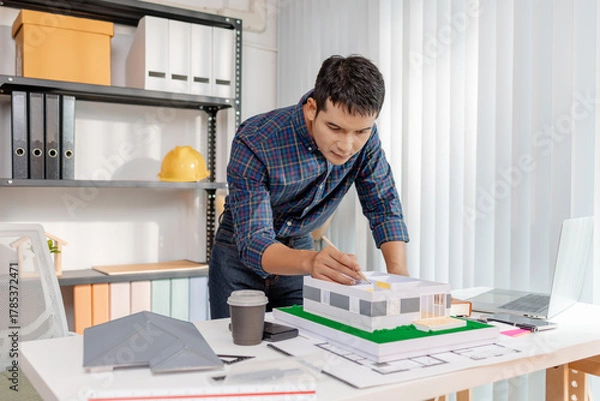 Fototapeta A young architect works with building plans and a house model in the office, reviewing blueprints, marking details, and analyzing design structures with focus and creativity.