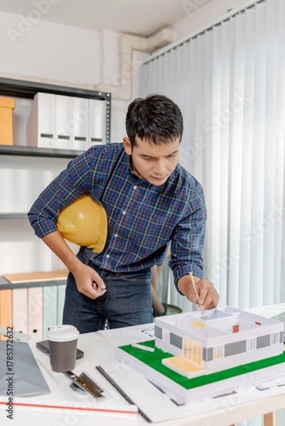 Fototapeta A young architect works with building plans and a house model in the office, reviewing blueprints, marking details, and analyzing design structures with focus and creativity.