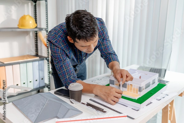 Fototapeta A young architect works with building plans and a house model in the office, reviewing blueprints, marking details, and analyzing design structures with focus and creativity.