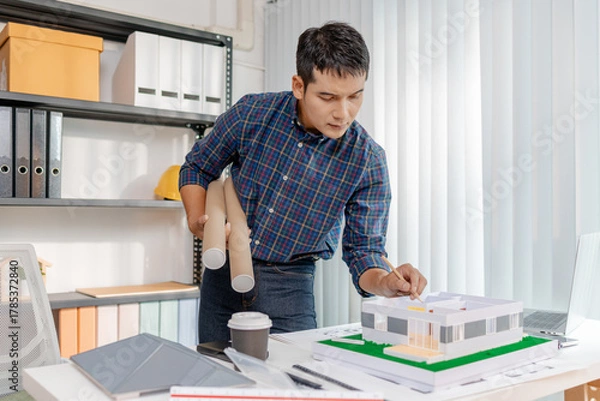 Fototapeta A young architect is working at his desk, reviewing blueprints, using a calculator, and checking a house model. He focuses on precision and detail while planning construction projects.