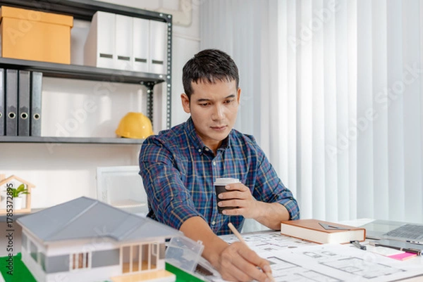 Fototapeta A young architect is working at his desk, reviewing blueprints, using a calculator, and checking a house model. He focuses on precision and detail while planning construction projects.
