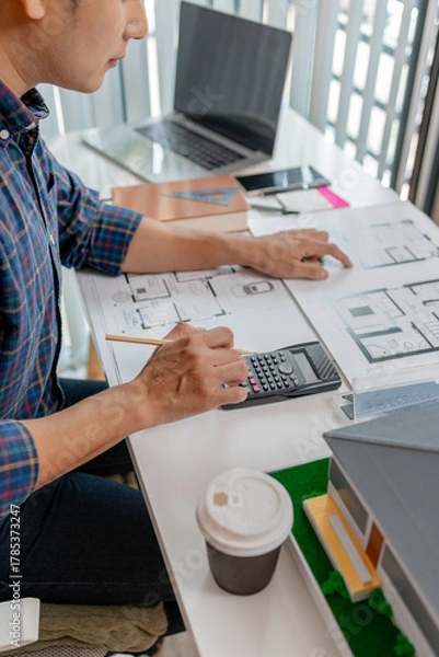 Fototapeta A young architect is working at his desk, reviewing blueprints, using a calculator, and checking a house model. He focuses on precision and detail while planning construction projects.