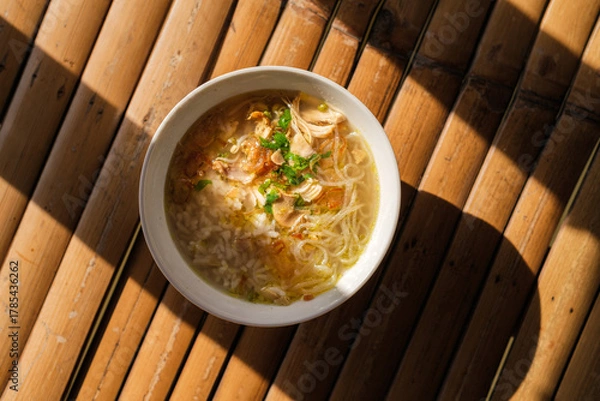 Fototapeta Overhead shot of nusantara indonesian food calles soto filled with chicken noodle soup in a white bowl, placed on a bamboo mat with a shadow.