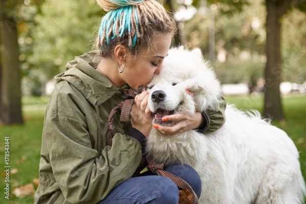 Fototapeta portrait of a young woman in a green coat and dreadlocks, kissing with his snow-white Samoyed dog in the autumn Park