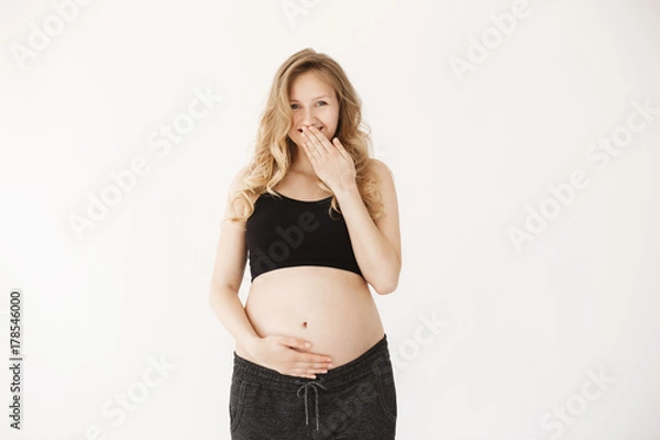 Fototapeta Close up of beautiful funny pregnant woman with blond curly hair in comfy sport clothes against white background in studio, closing mouth with hand, holding belly with happy face expression