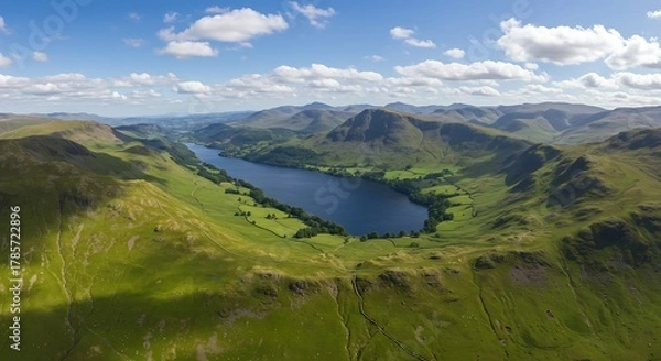 Fototapeta Aerial vista of a serene lake nestled amidst rolling green hills and majestic mountain ranges under a blue sky