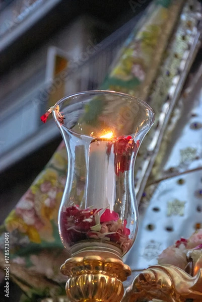 Fototapeta Lit candle in a glass holder with flower petals, captured during a religious procession in Granada, with the Virgin's cloak in the background. faith, ceremony