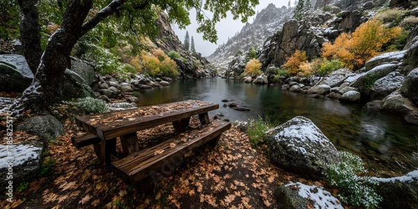 Fototapeta Peaceful mountain river scene with a wooden picnic table, autumn leaves, and a dusting of snow, capturing the quiet beauty of a changing season in a serene forest landscape.
