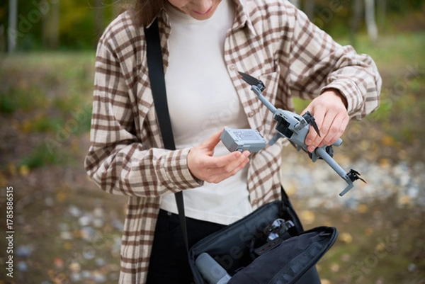 Fototapeta A young person in a checked shirt inspects a drone battery while getting ready to fly the drone in a beautiful forest setting. Vibrant autumn leaves surround the area.