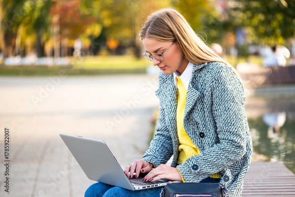 Obraz Portrait of confident businesswoman with eyeglasses  in coat using laptop while sitting on a park bench in the city during sunny autumn day.
