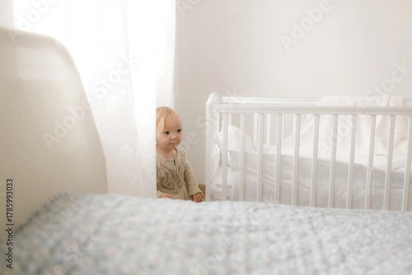 Fototapeta cute smiling healthy baby girl in beige bodysuit is standing near the crib at home in the nursery with teddy bears on white bedding, the child is learning to walk