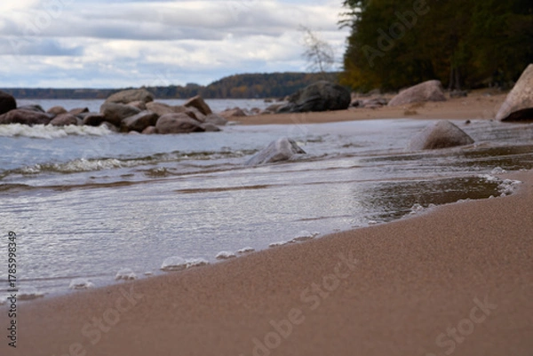 Fototapeta Low angle view of small waves reaching sandy beach with rocks and trees.