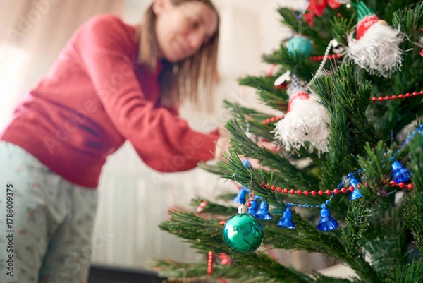 Fototapeta Woman enjoys the Christmas spirit and smiles while hanging bright and colorful ornaments on the tree