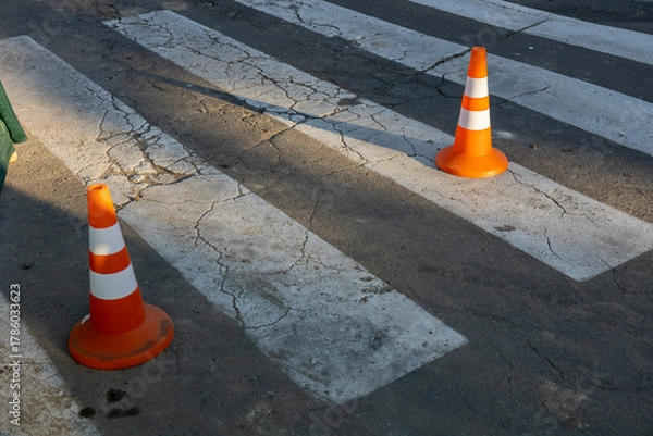 Fototapeta Two orange traffic cones are placed next to a pedestrian crosswalk on a cracked pavement as shadows stretch in the afternoon light