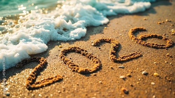 Fototapeta Close-up of "2026" written in golden sand on a sunlit beach with foamy ocean waves