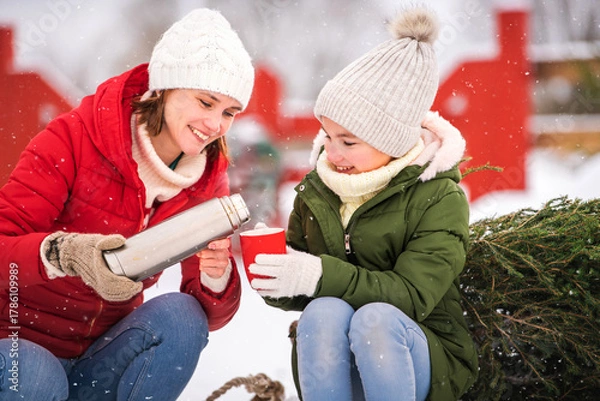 Obraz Happy woman pouring hot drink from thermos to child in red mug on snowy winter day.