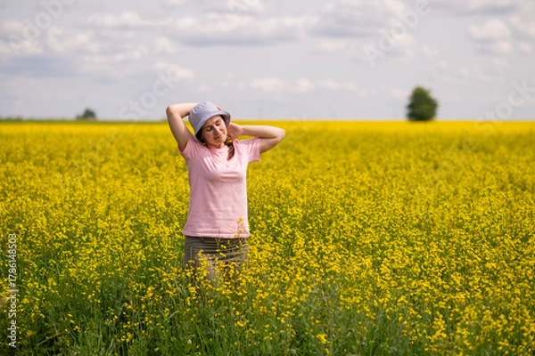 Fototapeta Woman in a bucket hat stands in yellow rapeseed field, enjoying the warmth and peace of a sunny summer day