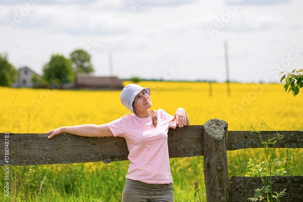 Fototapeta Young woman wearing a bucket hat leaning on an old wooden fence enjoying a warm summer day against a rustic farmstead surrounded by blooming rapeseed fields