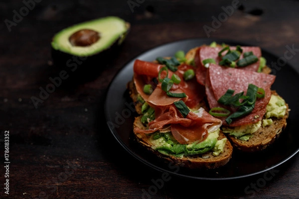 Obraz Close-up of avocado toast with salami, prosciutto, and green onions on a dark plate, healthy food and a satisfying meal.