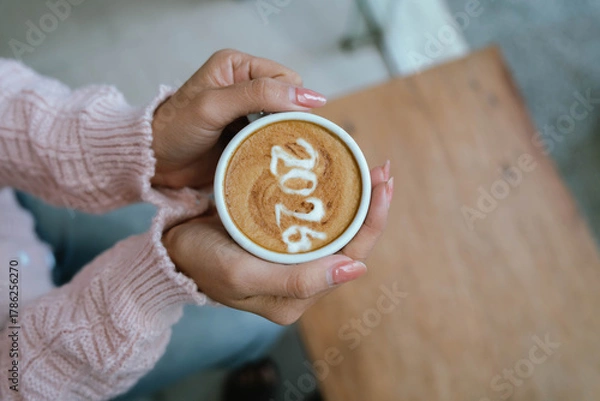 Fototapeta A person holding a cup of coffee with latte art showing the number "2026," wearing a pink sweater and relaxing indoors.