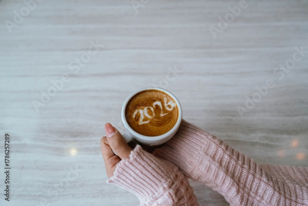 Fototapeta A person holding a cup of coffee with latte art showing the number "2026," wearing a pink sweater and relaxing indoors.