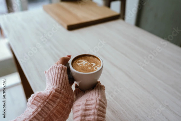 Fototapeta A person holding a cup of coffee with latte art showing the number "2026," wearing a pink sweater and relaxing indoors.