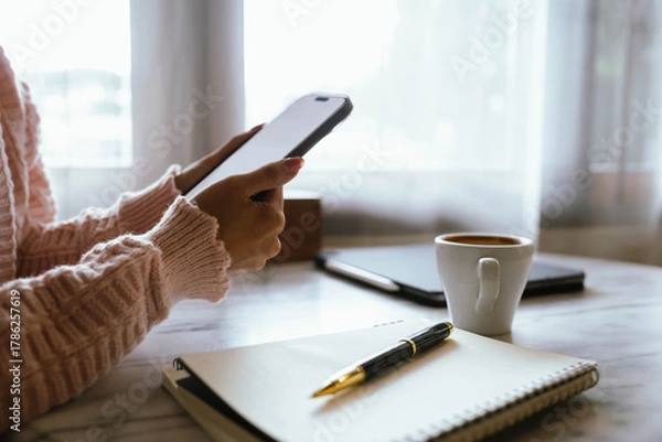 Fototapeta Woman using smartphone at cozy workspace with laptop, notebook, and coffee cup on marble table in soft daylight atmosphere.