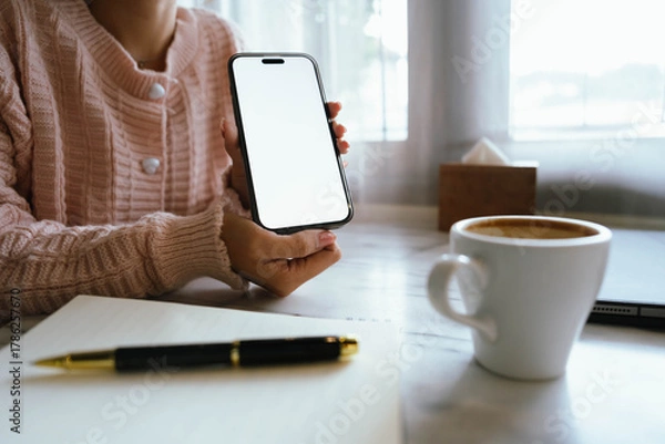 Fototapeta Woman using smartphone at cozy workspace with laptop, notebook, and coffee cup on marble table in soft daylight atmosphere.
