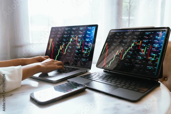 Fototapeta A person holding a smartphone showing stock market data with trading charts displayed on laptop screens in the background.